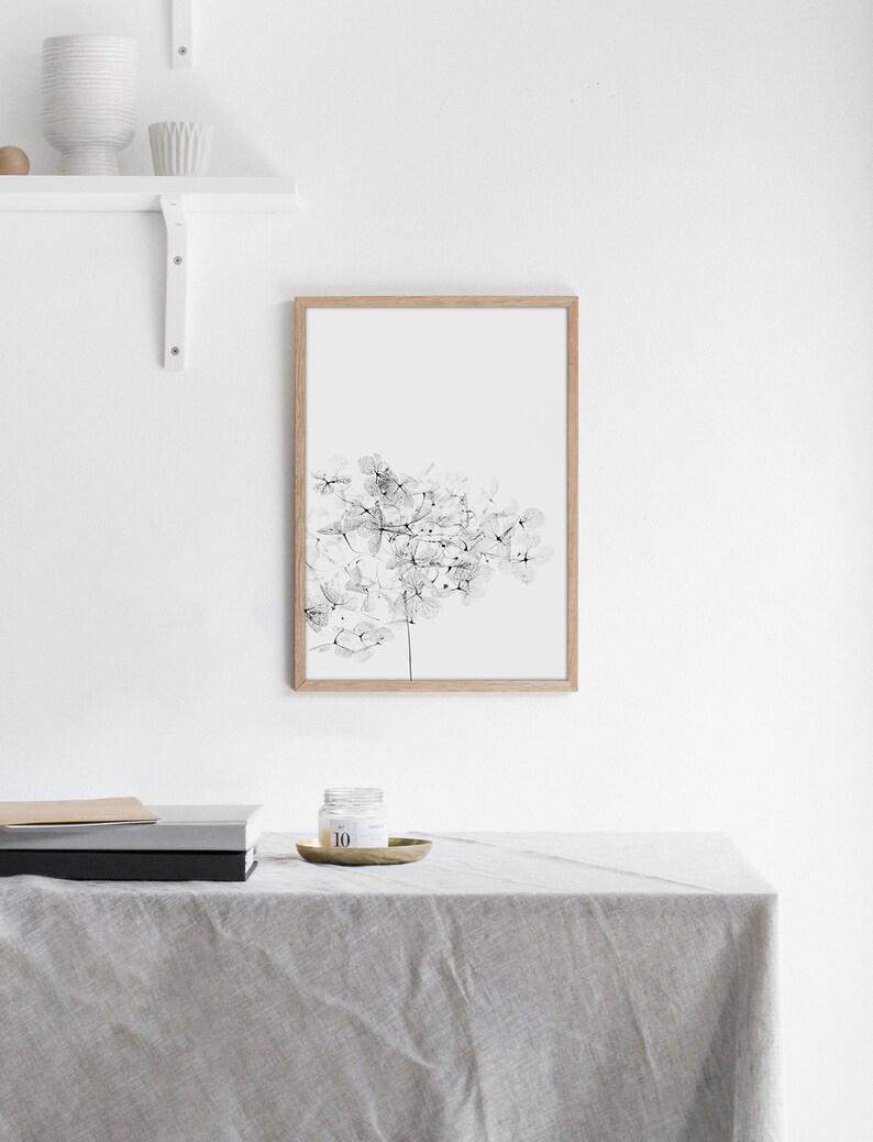 May include: A black and white line art print of a flower in a wooden frame. The print is hanging on a white wall above a table with a candle and books.
