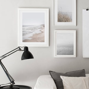 Puede incluir: Tres fotograf&iacute;as enmarcadas de paisajes marinos y costeros en un ambiente de sala de estar minimalista. Las fotos est&aacute;n en marcos blancos y presentan una playa con olas, un mar brumoso y un campo de hierba alta.