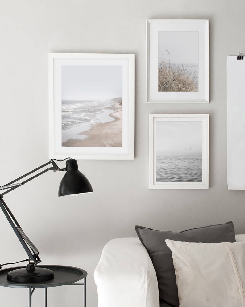 Puede incluir: Tres fotograf&iacute;as enmarcadas de paisajes marinos y costeros en un ambiente de sala de estar minimalista. Las fotos est&aacute;n en marcos blancos y presentan una playa con olas, un mar brumoso y un campo de hierba alta.