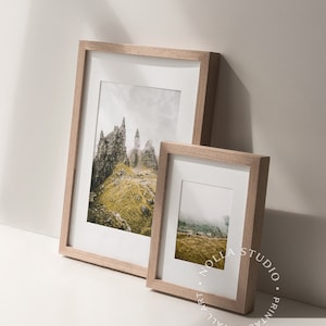 May include: Two framed landscape photographs with a light wood frame. The larger photo shows a rocky mountain range with a cloudy sky. The smaller photo shows a foggy mountain range with a green field in the foreground.