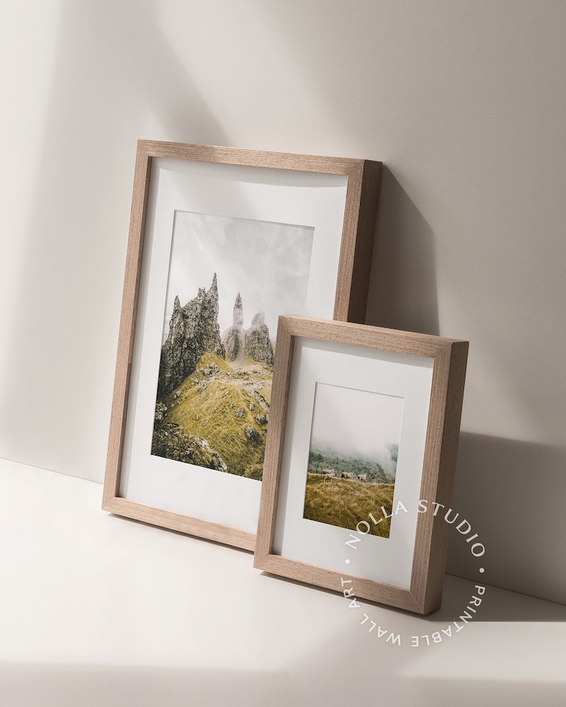 May include: Two framed landscape photographs with a light wood frame. The larger photo shows a rocky mountain range with a cloudy sky. The smaller photo shows a foggy mountain range with a green field in the foreground.