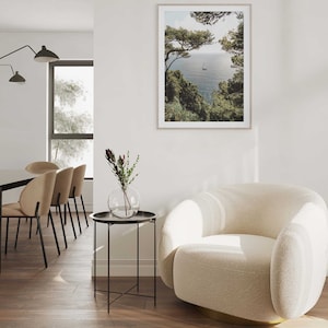 K&ouml;nnte beinhalten: Ein wei&szlig;er, flauschiger Sessel mit goldener Basis steht in einem Wohnzimmer mit einem Beistelltisch und einem gerahmten Foto einer Meereslandschaft. Das Foto zeigt ein Segelboot auf dem Wasser mit gr&uuml;nen B&auml;umen im Vordergrund.