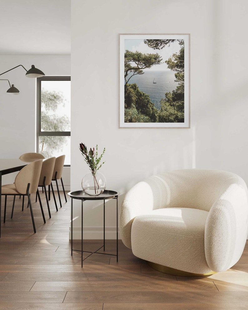 K&ouml;nnte beinhalten: Ein wei&szlig;er, flauschiger Sessel mit goldener Basis steht in einem Wohnzimmer mit einem Beistelltisch und einem gerahmten Foto einer Meereslandschaft. Das Foto zeigt ein Segelboot auf dem Wasser mit gr&uuml;nen B&auml;umen im Vordergrund.
