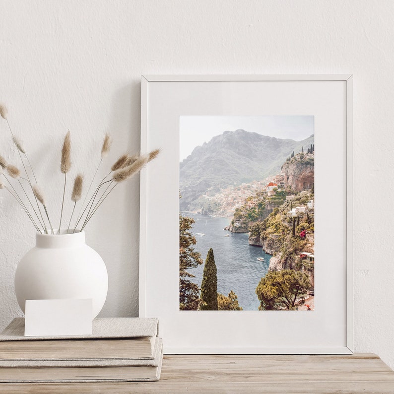 May include: A framed photograph of a coastal town nestled into a hillside overlooking a blue body of water. The town is surrounded by lush green trees and the water is dotted with boats.
