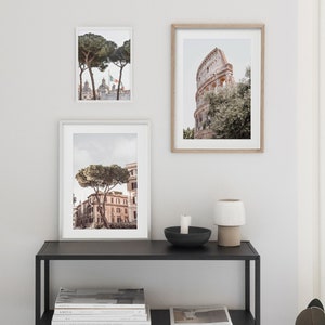 May include: Three framed photographs of Rome, Italy, with a black metal shelf with two shelves and a white ceramic lamp on top. The top shelf has a black candle holder with a white candle. The bottom shelf has a stack of magazines and a newspaper.