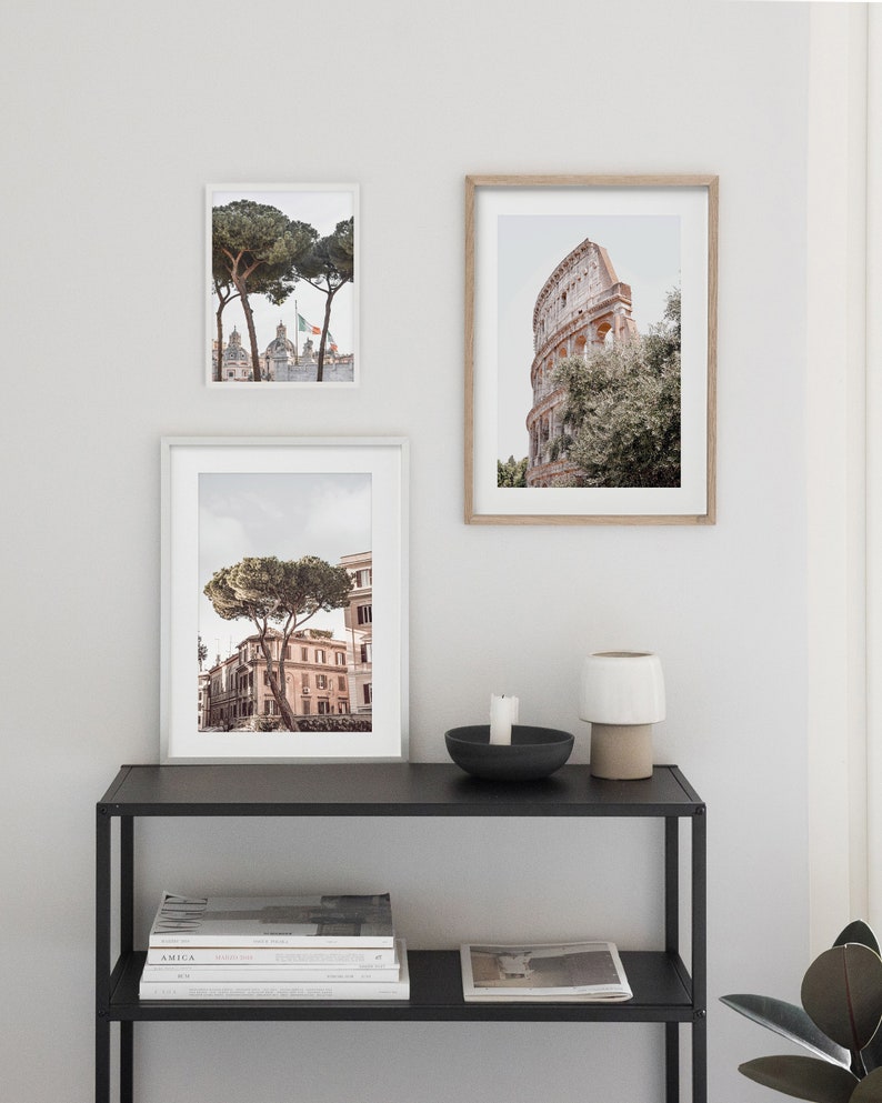 May include: Three framed photographs of Rome, Italy, with a black metal shelf with two shelves and a white ceramic lamp on top. The top shelf has a black candle holder with a white candle. The bottom shelf has a stack of magazines and a newspaper.