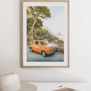 May include: A vintage orange Fiat car driving on a winding road with a lush green forest and a blue ocean in the background.