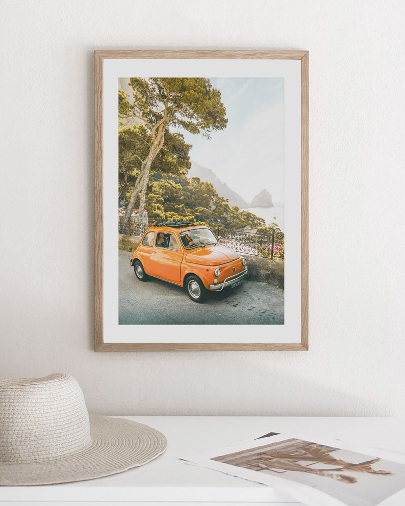 May include: A vintage orange Fiat car driving on a winding road with a lush green forest and a blue ocean in the background.