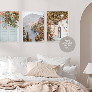 May include: Three framed prints of Italian coastal scenes. The first print shows a blue door with a white frame and a vine with pink flowers. The second print shows a view of a coastal town with a blue sea and green hills. The third print shows a white building with a staircase and green plants.