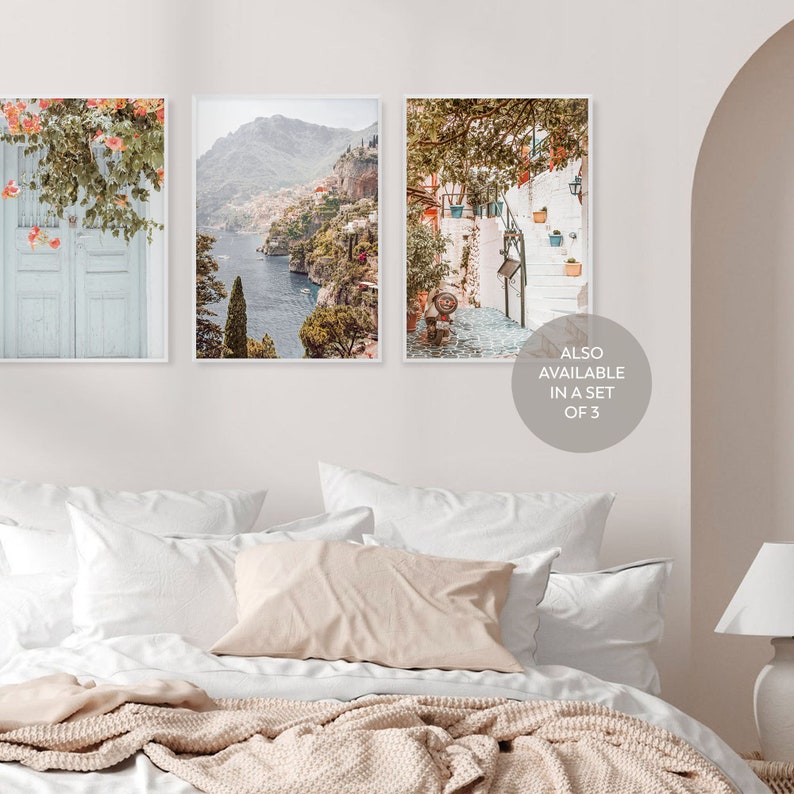 May include: Three framed prints of Italian coastal scenes. The first print shows a blue door with a white frame and a vine with pink flowers. The second print shows a view of a coastal town with a blue sea and green hills. The third print shows a white building with a staircase and green plants.