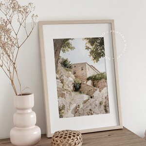 May include: A framed print of a stone building with a staircase leading up to it. The building is surrounded by trees and the sky is visible in the background. The print is in a light wood frame and is sitting on a wooden table.