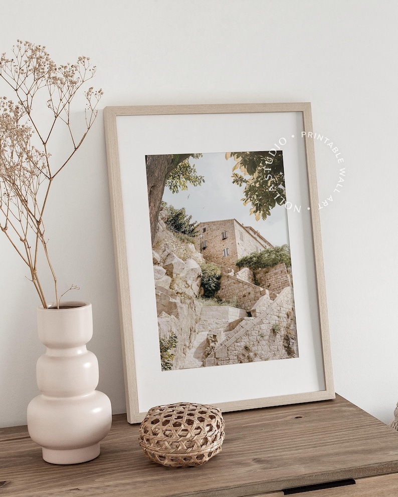 May include: A framed print of a stone building with a staircase leading up to it. The building is surrounded by trees and the sky is visible in the background. The print is in a light wood frame and is sitting on a wooden table.