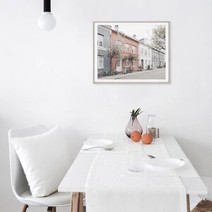 May include: A white dining table with a white tablecloth and a white pillow on a white chair. There is a framed print of a row of colorful houses with a tree in front of them hanging on the wall. There are two oranges and a vase with flowers on the table.
