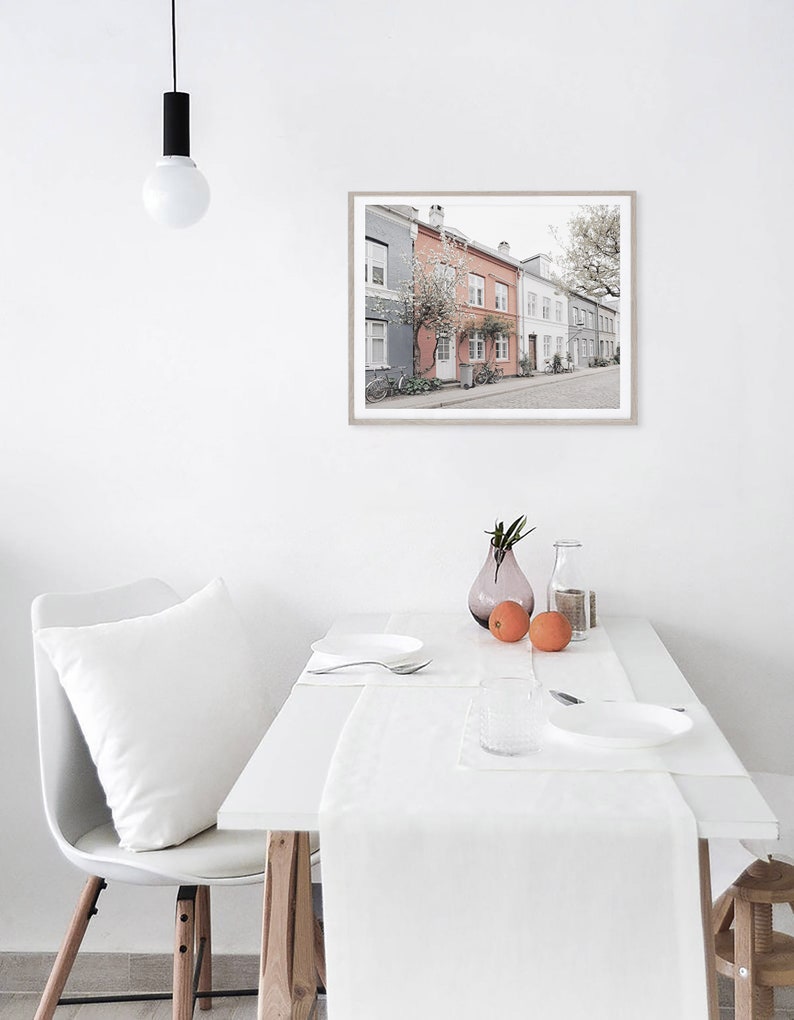 May include: A white dining table with a white tablecloth and a white pillow on a white chair. There is a framed print of a row of colorful houses with a tree in front of them hanging on the wall. There are two oranges and a vase with flowers on the table.