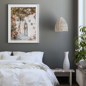 May include: A white framed photograph of a narrow, white-washed street with steps leading up to a building. The street is lined with potted plants and trees. The photo is hanging on a gray wall above a white bed with white bedding.