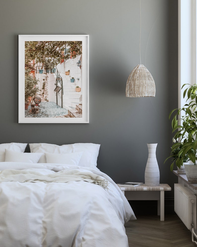May include: A white framed photograph of a narrow, white-washed street with steps leading up to a building. The street is lined with potted plants and trees. The photo is hanging on a gray wall above a white bed with white bedding.