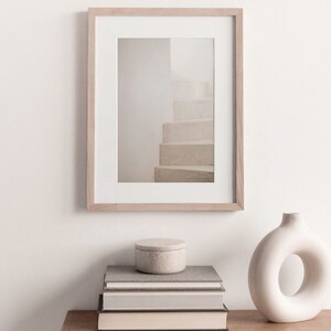 Peut inclure: Une photographie encadr&eacute;e d'un escalier blanc avec un cadre en bois brun clair. La photo est pos&eacute;e sur une table en bois avec une pile de livres et un vase en c&eacute;ramique blanc.