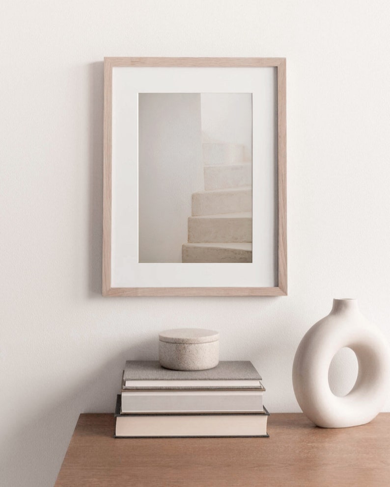 Peut inclure: Une photographie encadr&eacute;e d'un escalier blanc avec un cadre en bois brun clair. La photo est pos&eacute;e sur une table en bois avec une pile de livres et un vase en c&eacute;ramique blanc.