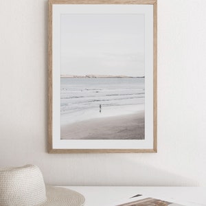 Puede incluir: Una fotograf&iacute;a enmarcada de una escena de playa con una persona caminando en el agua. La playa es arenosa y el agua es de color azul claro. El cielo es de color gris claro.