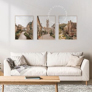 May include: Set of three framed prints featuring scenic views of a European city with cobblestone streets, stone buildings, and a church steeple. The prints are in a rustic style with a muted color palette.