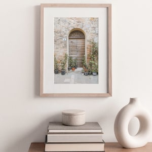 Pode incluir: Uma fotografia emoldurada de um edif&iacute;cio de pedra r&uacute;stico com uma porta de madeira e plantas em vasos. A porta tem um arco arredondado e &eacute; pintada de uma cor marrom clara. As plantas s&atilde;o verdes e frondosas, e est&atilde;o dispostas em uma fileira na frente da porta.
