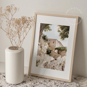 May include: A framed print of a stone building with steps leading up to it. The building is surrounded by trees and greenery. The print is in a light wood frame.