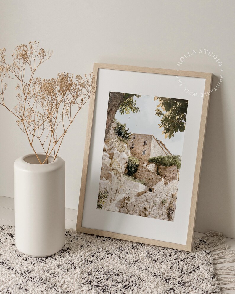 May include: A framed print of a stone building with steps leading up to it. The building is surrounded by trees and greenery. The print is in a light wood frame.