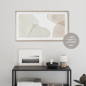 Op de afbeelding: Een moderne woonkamer met een ingelijst abstract schilderij aan de muur boven een zwart rek. Het schilderij toont zachte groene en bruine aquarelvormen op een witte achtergrond. Het rek bevat een ingelijste zwart-witfoto, een zwarte kom met een witte kaars en een witte keramische lamp met een bruine voet. De tekst "TURN YOUR TV INTO ART" is zichtbaar in de rechterbenedenhoek van de afbeelding.