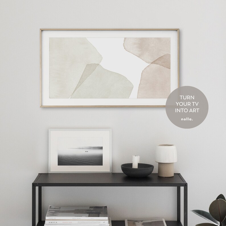 Op de afbeelding: Een moderne woonkamer met een ingelijst abstract schilderij aan de muur boven een zwart rek. Het schilderij toont zachte groene en bruine aquarelvormen op een witte achtergrond. Het rek bevat een ingelijste zwart-witfoto, een zwarte kom met een witte kaars en een witte keramische lamp met een bruine voet. De tekst "TURN YOUR TV INTO ART" is zichtbaar in de rechterbenedenhoek van de afbeelding.