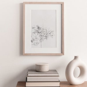 May include: A black and white print of a delicate flower arrangement in a light wood frame, displayed on a wooden shelf with a stack of books and a white ceramic vase.