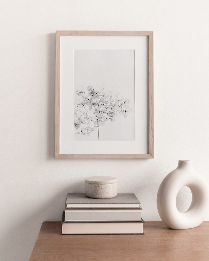 May include: A black and white print of a delicate flower arrangement in a light wood frame, displayed on a wooden shelf with a stack of books and a white ceramic vase.