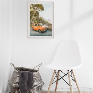 May include: A framed photograph of an orange vintage car parked on a winding road overlooking a blue ocean. The car is surrounded by lush green trees and the sky is a light blue.