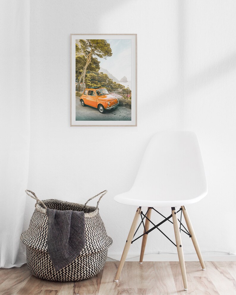 May include: A framed photograph of an orange vintage car parked on a winding road overlooking a blue ocean. The car is surrounded by lush green trees and the sky is a light blue.