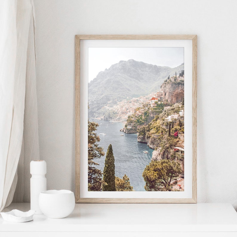 May include: A framed photograph of a coastal landscape with a view of a town built into the side of a mountain. The town is nestled between the mountain and the sea. The water is a deep blue and the sky is a light blue. The photo is framed in a light wood frame.