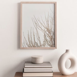 May include: A framed print of a minimalist nature scene with dried branches against a white background. The print is displayed on a wooden table with a stack of books and a white ceramic vase.