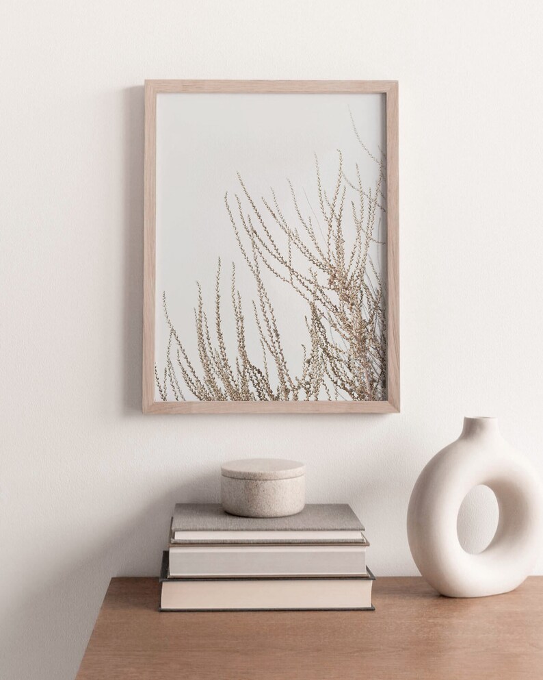 May include: A framed print of a minimalist nature scene with dried branches against a white background. The print is displayed on a wooden table with a stack of books and a white ceramic vase.