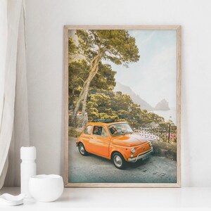 May include: A vintage orange Fiat car driving on a winding road through a lush green forest with a view of the ocean in the background.