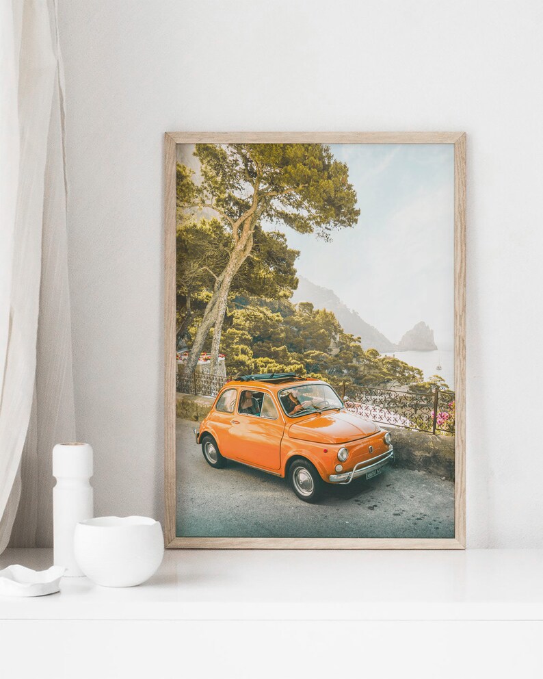 May include: A vintage orange Fiat car driving on a winding road through a lush green forest with a view of the ocean in the background.