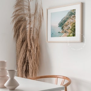 May include: A white table with a wooden chair sits in front of a white wall with a framed print of a coastal town. A tall vase with dried pampas grass is in the corner of the room.