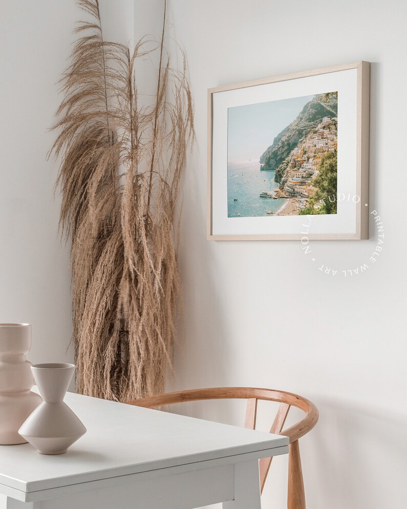 May include: A white table with a wooden chair sits in front of a white wall with a framed print of a coastal town. A tall vase with dried pampas grass is in the corner of the room.