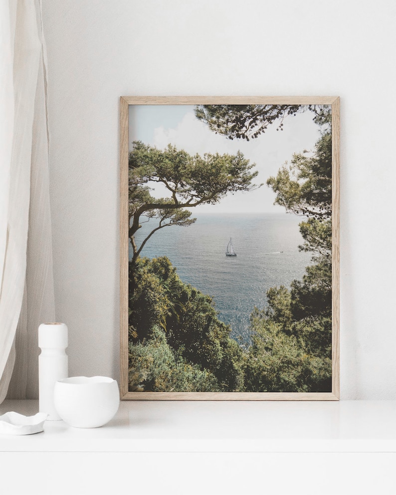 K&ouml;nnte beinhalten: Ein gerahmtes Foto einer Meereslandschaft mit einem Segelboot in der Ferne. Die Szene wird von &uuml;ppigen gr&uuml;nen B&auml;umen auf beiden Seiten des Bildes eingerahmt.