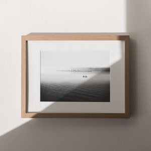 May include: A black and white photograph of a lake with a small boat in the distance. The lake is covered in fog and the sky is overcast. The photo is framed in a light wood frame.
