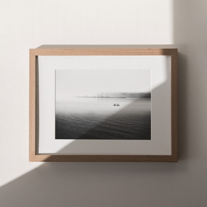 May include: A black and white photograph of a lake with a small boat in the distance. The lake is covered in fog and the sky is overcast. The photo is framed in a light wood frame.