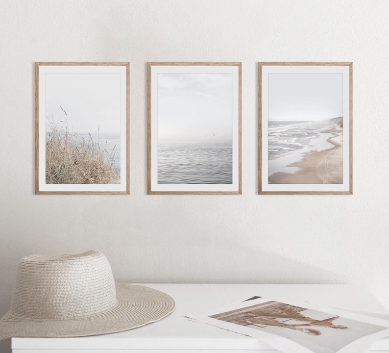 Puede incluir: Tres impresiones enmarcadas de paisajes marinos en un estilo minimalista. Las impresiones presentan una playa con olas, un paisaje marino con un p&aacute;jaro volando por encima y un primer plano de hierbas altas.