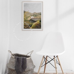 May include: A framed photograph of a train traveling through a green valley with a stone bridge. The train is emitting steam. The photograph is hanging on a white wall with a white chair and a woven basket in the foreground.