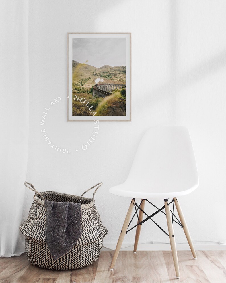 May include: A framed photograph of a train traveling through a green valley with a stone bridge. The train is emitting steam. The photograph is hanging on a white wall with a white chair and a woven basket in the foreground.