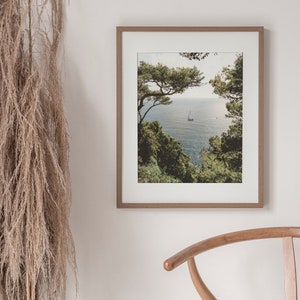 K&ouml;nnte beinhalten: Ein gerahmtes Foto einer Meereslandschaft mit einem Segelboot in der Ferne, eingerahmt von &uuml;ppigen gr&uuml;nen B&auml;umen. Das Foto ist in einem hellen Holzrahmen.