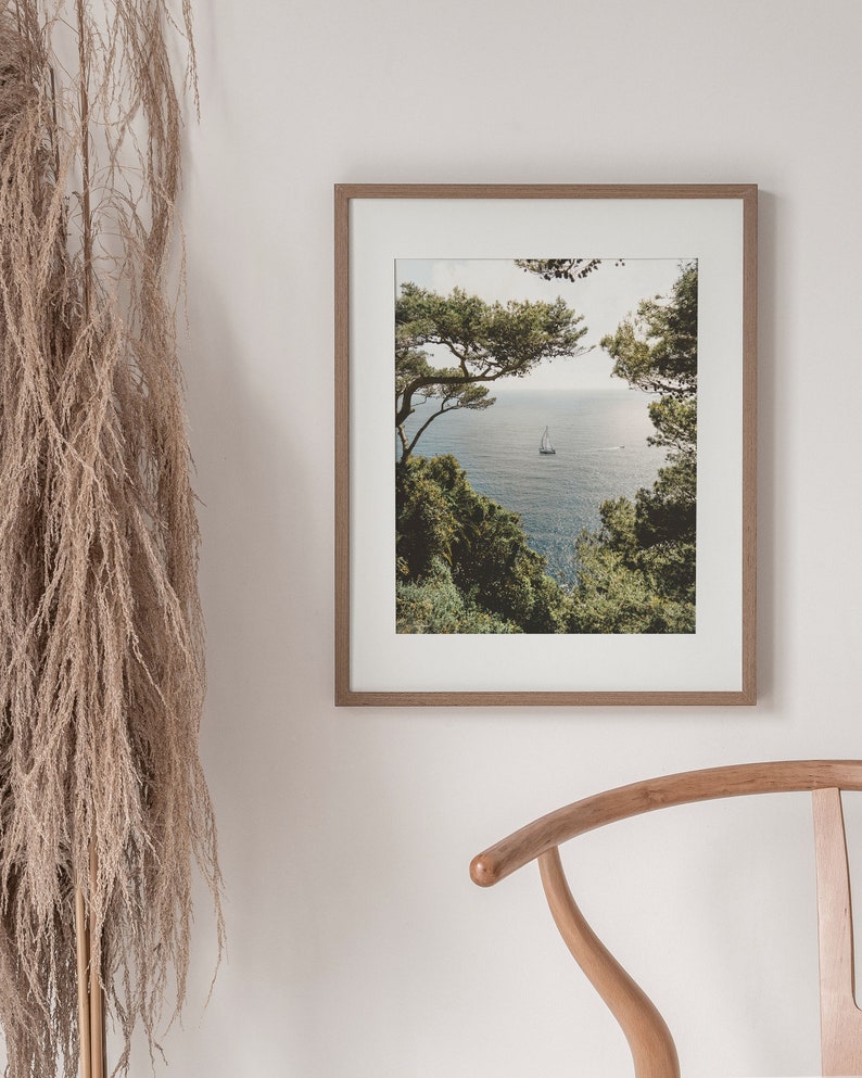K&ouml;nnte beinhalten: Ein gerahmtes Foto einer Meereslandschaft mit einem Segelboot in der Ferne, eingerahmt von &uuml;ppigen gr&uuml;nen B&auml;umen. Das Foto ist in einem hellen Holzrahmen.