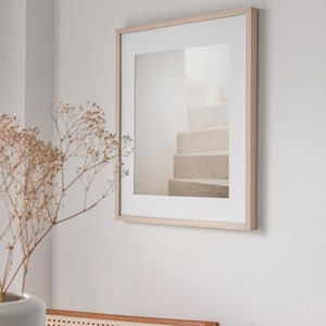 Peut inclure: Une photographie encadr&eacute;e d'un escalier blanc avec un cadre en bois clair. La photographie est accroch&eacute;e &agrave; un mur blanc.