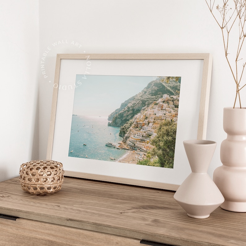 May include: A framed photograph of a coastal town with colorful buildings on a hillside overlooking a blue ocean. The photo is displayed on a wooden surface with decorative vases and a woven basket.